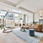 Helles Wohnzimmer mit Ledersofa, Couchtisch, blauem Teppich und Hängesessel, kombiniert mit großen Fenstern und natürlicher Einrichtung.